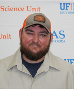 Man with brown beard wearing a tan collared shirt and ball cap smiles at the camera.