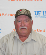 Man with gray mustache wearing a tan collared shirt and camouflage ball cap smiles at the camera.