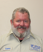 A man with short gray hair and a fluffy gray beard smiles at the camera wearing a tan shirt.